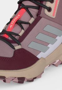 Gros plan sur une chaussure de randonnée violette et beige avec des lacets noirs, des bandes grises réfléchissantes, une étiquette rouge « TERREX » et une semelle robuste.
