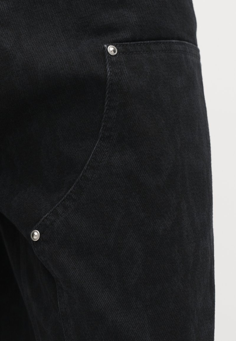 Nahaufnahme einer schwarzen Denim-Hose mit Fronttasche und silbernen Nieten auf hellem Hintergrund.