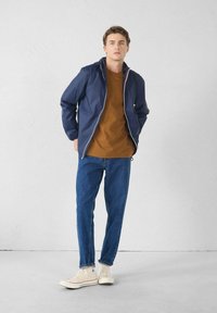Wasserabweisende marineblaue Jacke mit Kapuze, Frontreißverschluss und kontrastierenden pinkfarbenen Details. Kombiniert mit einem braunen T-Shirt und blauen Jeans.