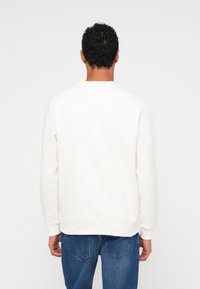 Sudadera blanca con cuello redondo, puños y dobladillo acanalados; combinada con jeans azules, con detalles de costura visibles en la prenda.