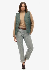 Zielona pikowana kamizelka z przednią kieszenią, beżowy golf i spodnie w paski. Na sobie brązowy pleciony pasek i ciemne buty.