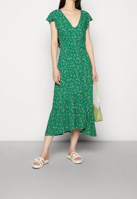 Grünes, florales Midi-Kleid mit kurzen Ärmeln, V-Ausschnitt und gerafftem Saum. Kombiniert mit weißen Sandalen und einer kleinen grünen Tasche.
