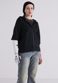 Crni dukserica s patentnim zatvaračem, opuštenog kroja i kratkim rukavima, u kombinaciji s svijetloplavim prugastim trapericama. Ima robotsku ruku.