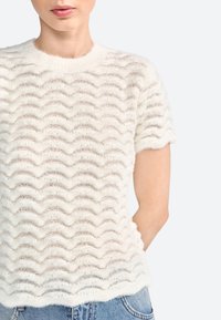 Hvid tekstureret kortærmet sweater med bølgemønstre og en blød, fuzzy finish. Kantene er let scalloperede, hvilket komplementerer designet.