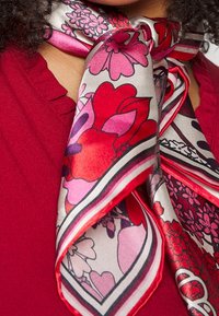 Écharpe en soie à motif floral éclatant en rouge, rose et blanc, nouée autour du cou sur un haut rouge, avec des cheveux foncés et bouclés visibles.