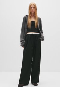 Šedý rebrovaný kardigán cez čierny crop top, spárovaný s voľnými čiernymi nohavicami a tmavými topánkami. Hladké textúry a uvoľnený strih.
