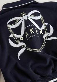 Tela azul marino con lazo de cinta blanca bordado y escudo con borde dorado, texto "Paris Baker by Ted Baker" en prenda texturizada.