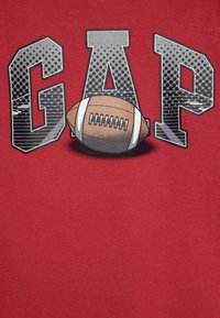 Camiseta roja con "GAP" en una fuente gris, en negrita y texturizada, y un balón de fútbol marrón colocado en la base de las letras. Material de algodón suave.