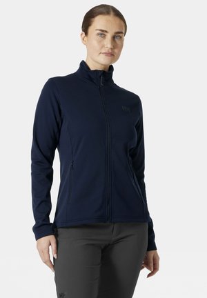 Mujer con chaqueta azul marino con cremallera y bolsillos laterales con cremallera, y pantalones gris oscuro, de pie frente a un fondo blanco.