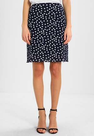 Jupe navy à pois fabriquée en tissu léger, avec des pois blancs, un ourlet en forme de feston et une silhouette ajustée, associée à des talons noirs à lanières.