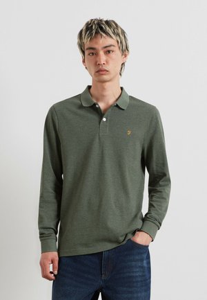 Giovane uomo con capelli biondi corti che indossa una polo verde a maniche lunghe e jeans blu, in piedi davanti a uno sfondo chiaro e uniforme.