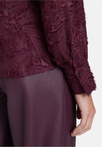 Primo piano di una mano accanto a una maglietta a maniche lunghe bordeaux con texture e pantaloni bordeaux lisci su uno sfondo semplice.