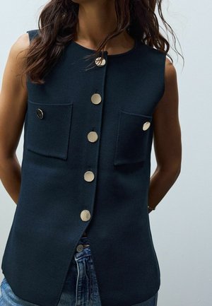 Vrouw draagt een mouwloos donkerblauw vest met grote ronde knopen en twee voorzakken, gecombineerd met een blauwe spijkerbroek.