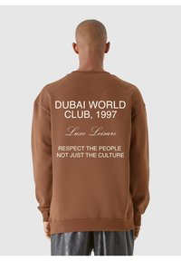 Felpa marrone con collo tondo, riportante sulla schiena testo bianco: "DUBAI WORLD CLUB, 1997," "Luxe Leisure," e una citazione in grassetto.