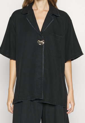 Mujer con camisa negra de manga corta y holgada, con cuello abierto en pico, un gran botón metálico y bolsillo en el pecho.