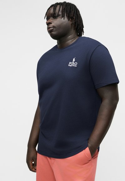 Mann mit Dreadlocks, der ein marineblaues Polo Ralph Lauren T-Shirt und korallenfarbene Shorts trägt, steht mit einer Hand in der Tasche vor einem schlichten Hintergrund.