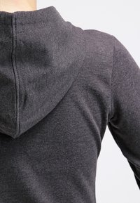 Personne portant un sweat à capuche gris foncé, montrant le dos et la capuche sur un fond clair uni.