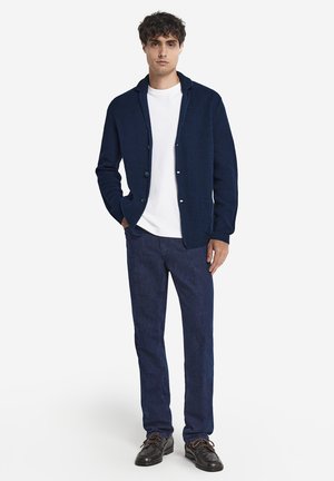 Marineblå strik cardigan med sjalskrave, udstyret med frontknapper og påsyede lommer, parret med slim-fit mørkeblå bukser og sorte sko.