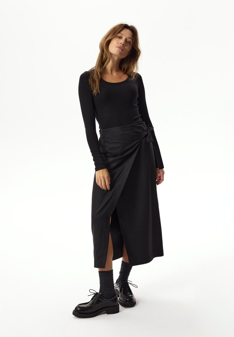 Czarny top z długim rękawem w połączeniu z czarną spódnicą w stylu owijanym, z wiązaniem i bocznymi rozcięciami, uzupełniony błyszczącymi czarnymi butami.