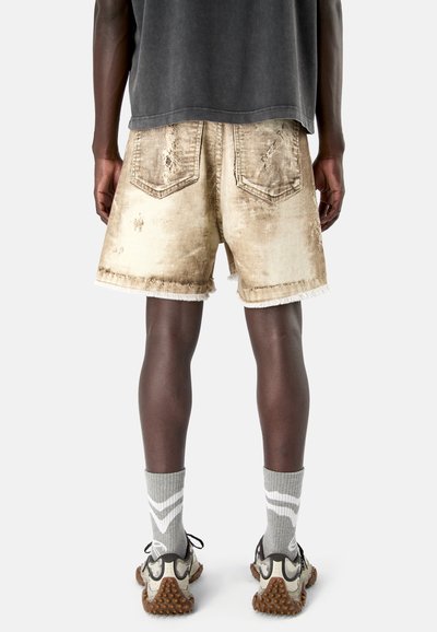 Person wearing beige distressed denim shorts, gray crew socks with white chevrons, and beige and black sneakers, shown from the back.