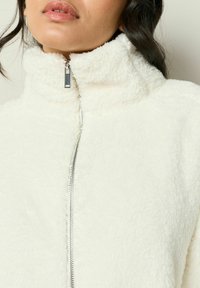 Hvid fleece zip-jakke med høj krave og sølvfarvet lynlås. Teksturen er blød og fyldig, med minimale synlige sømme.