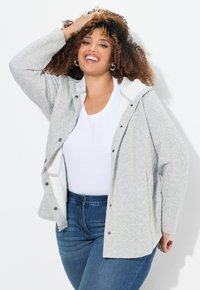Světlé šedé prošívané sako s bílou kapucí a patentkami, nošené přes bílou košili a modré džíny. Má volný střih a texturovanou látku.