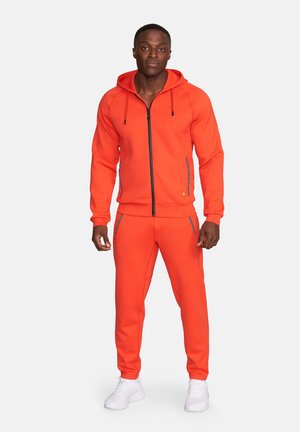 Mann steht und trägt eine leuchtend orangefarbene Kapuzenjacke mit Reißverschluss und passende Hosen sowie weiße Turnschuhe vor einem einfarbigen Hintergrund.