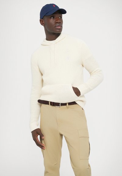 Krémovo sfarbená pletená mikina s kapucňou a predným vreckom, doplnená o khaki cargo nohavice a tmavomodrú čiapku s červeným logom.