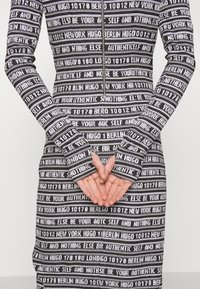 Robe à manches longues noire et blanche avec des rayures de texte horizontal comprenant "HUGO", "NEW YORK", "BERLIN" et "AUTHENTIC SELF", mains croisées dans le dos.