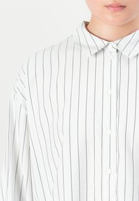 Nærbilde av en person som har på seg en hvit skjorte med tynne svarte vertikale striper og en klassisk krage.