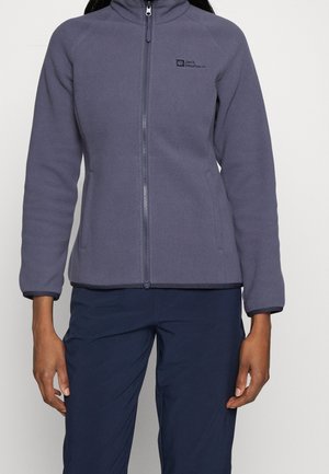 Personne portant une veste polaire grise zippée à manches longues et un pantalon bleu foncé, debout devant un fond uni.