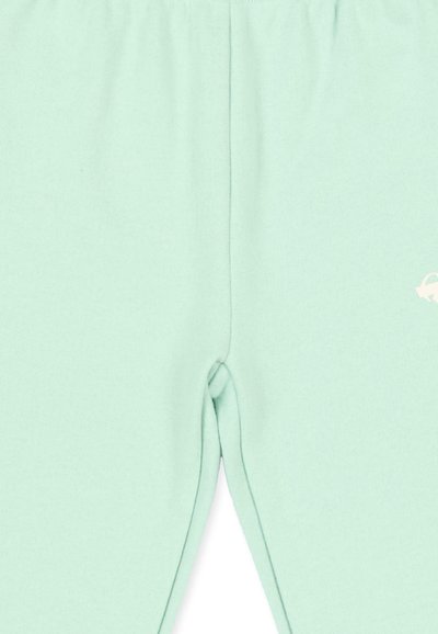 Pantalon vert clair avec une couture centrale et un petit logo blanc sur la partie supérieure droite de la cuisse, texture du tissu visible.