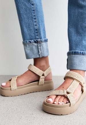 Sandales à plateforme beiges avec des brides tressées, une semelle épaisse en caoutchouc et une finition texturée. Portées avec un jean en denim retroussé.