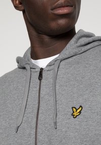 Grijze hoodie met ritssluiting, voorzien van een gestructureerde stof, verstelbare trekkoorden en een geel geborduurd arendlogo op de borst.