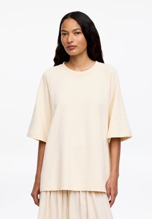 Donna con capelli lunghi e scuri che indossa un top a maniche corte color crema e una gonna abbinata su uno sfondo bianco semplice.