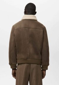 Giacca in shearling marrone con colletto in pelliccia, polsini e orlo a coste. Texture morbida e design strutturato con un semplice dettaglio della cucitura sul retro.