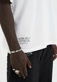 White cotton t-shirt featuring black printed logo "Don Ed Hardy designs" at the hem, worn with dark denim jeans and silver jewelry accents.