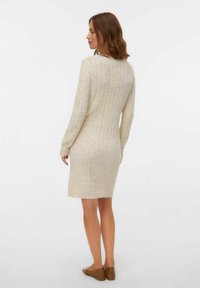 Naine, kellel on õlgadele ulatuvad pruunid juuksed, kannab beeži soonikkoes pika varrukaga kleiti ja pruune lame kingi, seistes seljaga pöördunult.