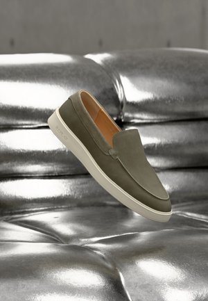 Scarpa slip-on verde oliva con suola bianca che fluttua sopra una superficie argentata lucida trapuntata.