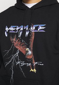 Sudadera negra con un gráfico de un águila roja y rayos. El texto dice "MENACE" en letras estilizadas azules y "Flying Free" en blanco.
