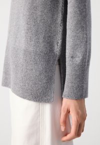 Hellgrauer, gestrickter Pulloverärmel und Saum mit seitlichem Schlitz über weißen Hosen, wobei eine entspannt an die Seite hängende Hand zu sehen ist.