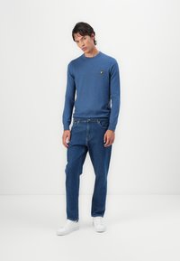 Pull bleu à col rond, en tissu tricot léger, associé à un jean droit en denim et des sneakers blanches. Petit logo jaune sur la poitrine.