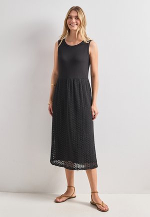 Femme debout et souriante portant une robe noire sans manches avec un haut uni et une jupe texturée, associée à des sandales marron et des bracelets simples.