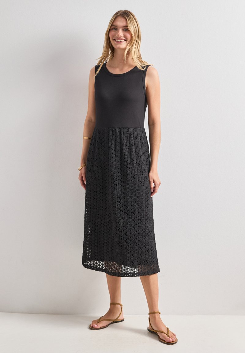 Femme debout et souriante portant une robe noire sans manches avec un haut uni et une jupe texturée, associée à des sandales marron et des bracelets simples.