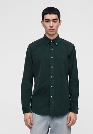 Jeune homme aux cheveux courts noirs porte une chemise verte foncée boutonnée et un jean bleu clair, debout devant un fond clair uni.