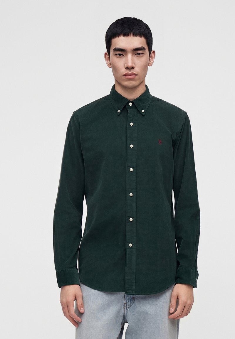 Jeune homme aux cheveux courts noirs porte une chemise verte foncée boutonnée et un jean bleu clair, debout devant un fond clair uni.