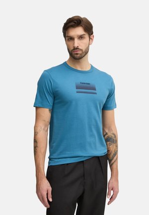 Camiseta de manga corta en color turquesa, fabricada en un material suave, que presenta una franja horizontal negra con el logo de "Calvin Klein" en el centro. Ajuste regular.