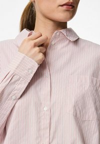 Vasker roosa nööbiga särk õhukeste vertikaalsete triipudega, krae ja rinna taskuga. Esitleb valgeid nööpe ja mansettide detailimist.