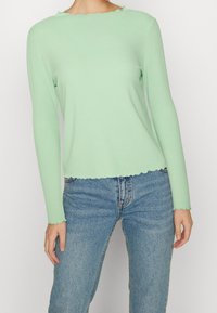 Světle zelený žebrovaný dlouhý rukáv s vykrojeným spodním lemem, zkombinovaný s modrými džínami. Tkanina vypadá měkce a přiléhavě.