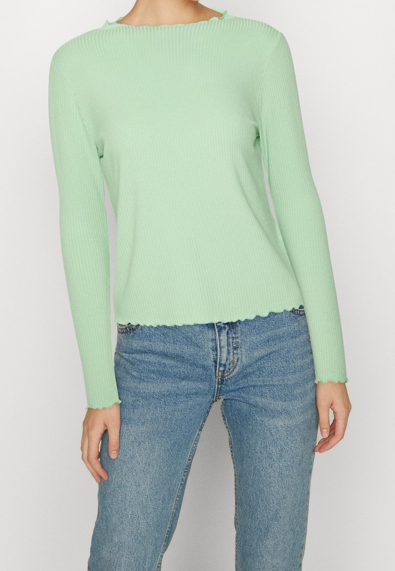 Světle zelený žebrovaný dlouhý rukáv s vykrojeným spodním lemem, zkombinovaný s modrými džínami. Tkanina vypadá měkce a přiléhavě.